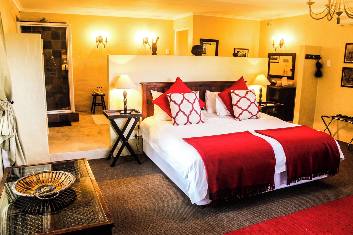 A hotel room with a large bed, two bedside tables, a glass coffee table, a red throw blanket, and a shower area.