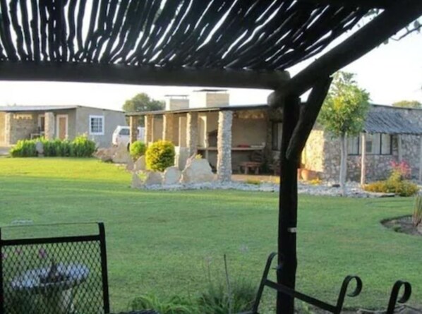 A covered outdoor area with a pergola structure, a stone building, and a well-maintained lawn. - Eldorado Guest House & Camping (Okaukuejo)