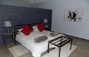 A bedroom with a bed, two nightstands, a lamp, a red pillow, a grey pillow, a wall art of a bull, and a wooden bench. - Eldorado Guest House & Camping (Okaukuejo)