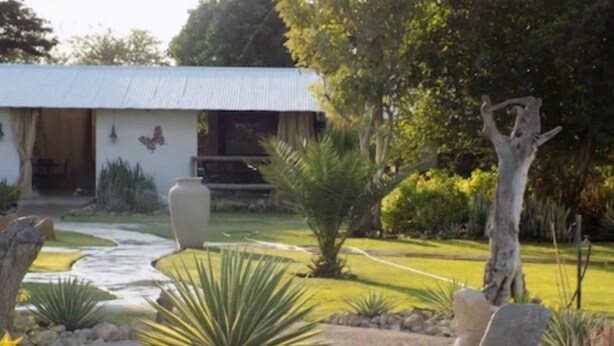 Un edificio rústico con techo de metal corrugado, rodeado por un jardín cuidado con diversas plantas y un sendero de piedra que conduce a la entrada.