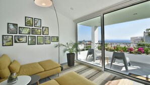 A modern living room with a yellow sofa, a small table, and a potted plant. There is a large window with a view of the sea and a balcony with outdoor furniture. - Coconut Villa (Alimos)