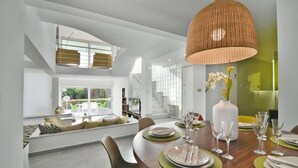 A modern dining area with a wooden table set for a meal, a large woven pendant light, and a view of a staircase and outdoor seating area. - Coconut Villa (Alimos)