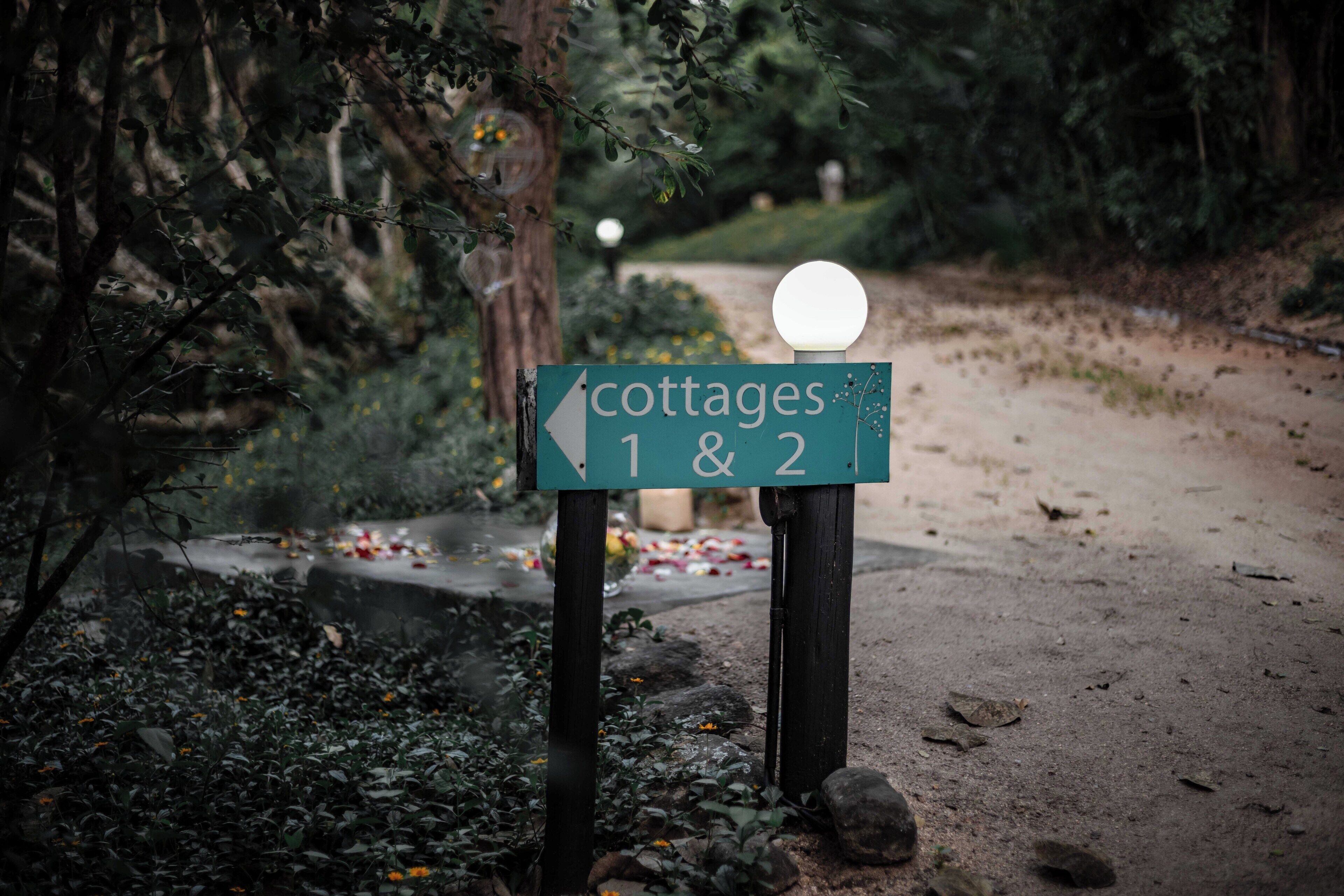 Señalización para 'cottages 1 & 2' con flecha indicativa, ubicada en un entorno natural y arbolado con sendero de tierra y farola.