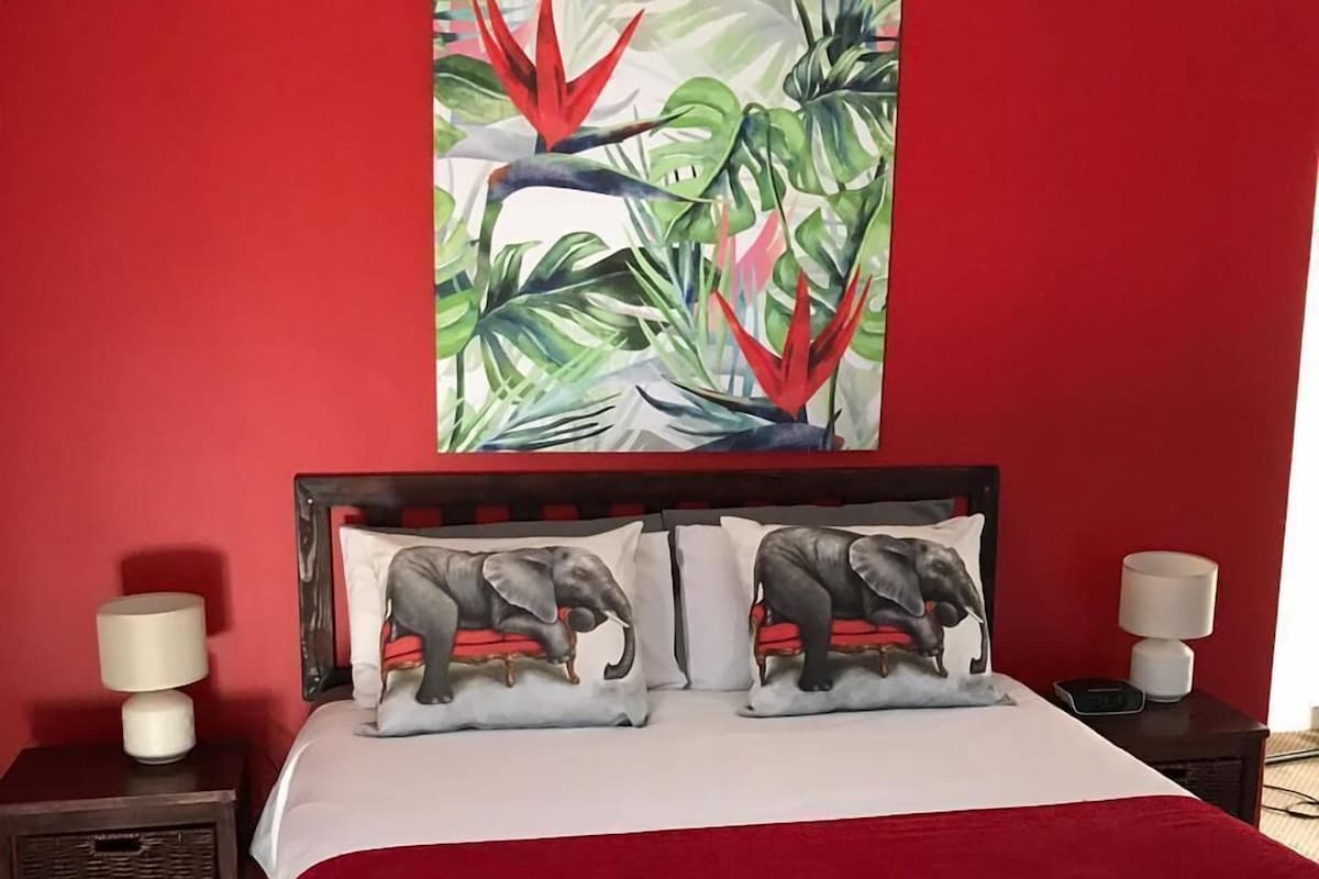 Un dormitorio con pared roja, un cuadro de una planta tropical grande, dos almohadones con motivos de elefantes, una colcha blanca y dos lámparas de noche.