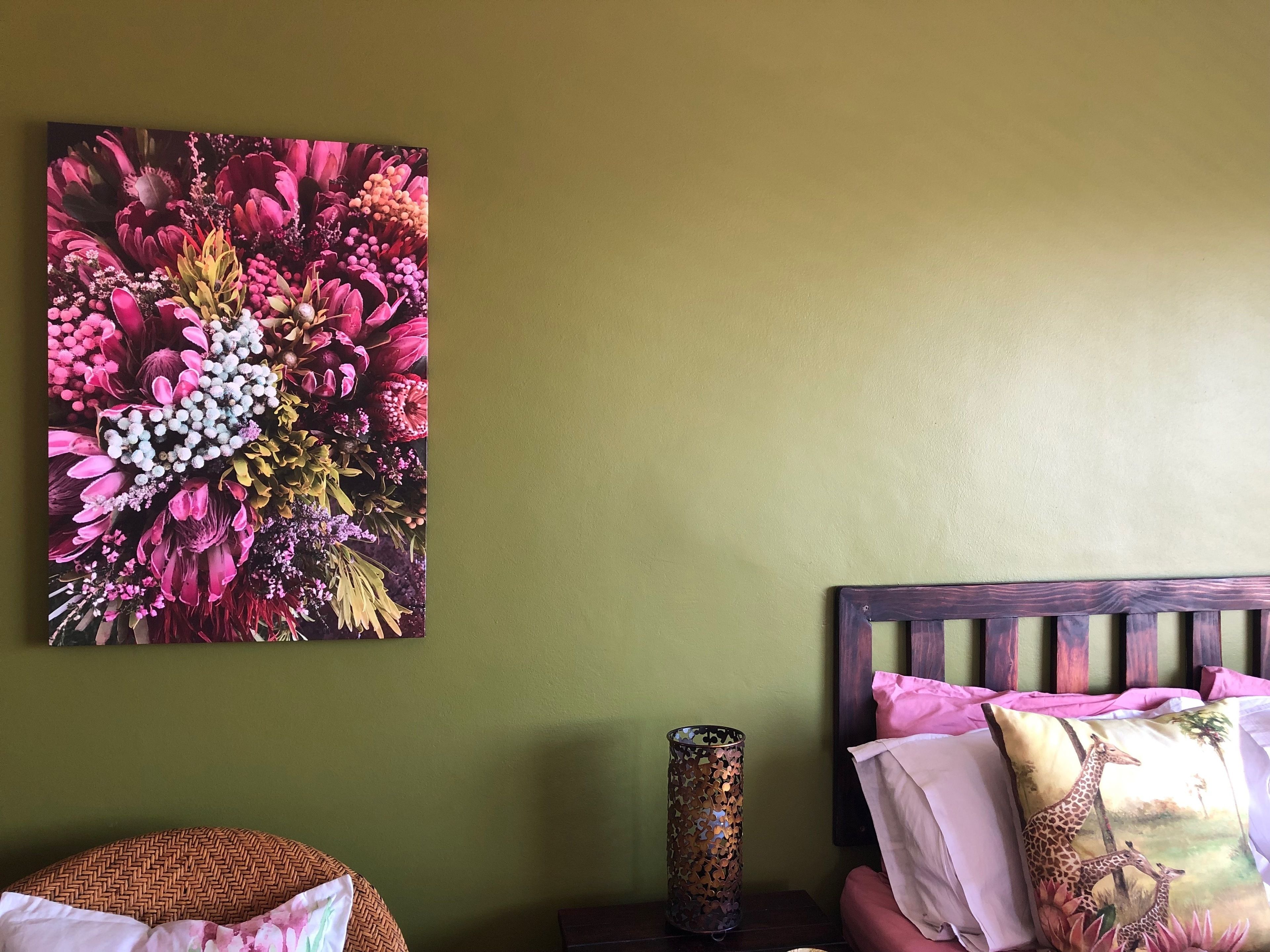 A room with a green wall, a floral painting, a decorative metal vase, a bed with a giraffe pillow, and a woven chair.