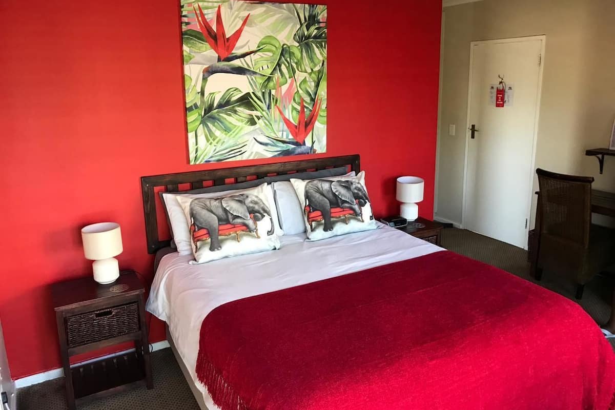 A bedroom with a red wall, a large painting, a bed with elephant pillows, two bedside lamps, and a red throw blanket.
