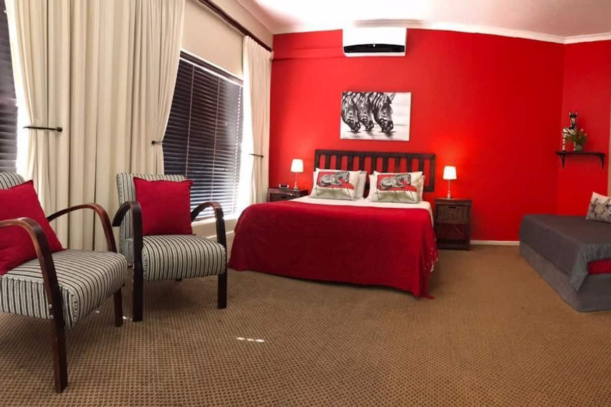 A hotel room with a red accent wall, a bed with a red bedspread, two striped chairs, a grey sofa, a small table with a lamp, and a television on the wall.