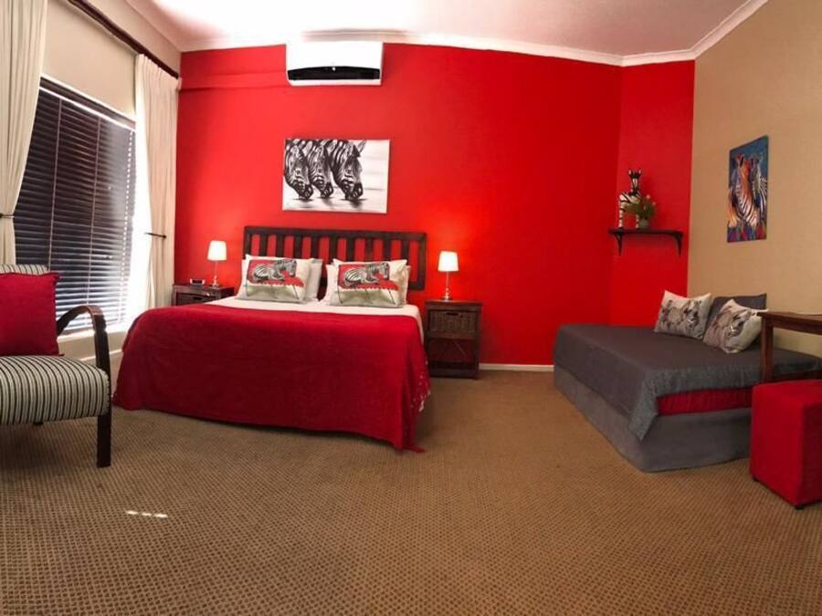 Habitación de hotel con una pared de acento roja, una cama con colcha roja, dos sillas a rayas, un sofá gris, una mesita con una lámpara y un televisor en la pared.