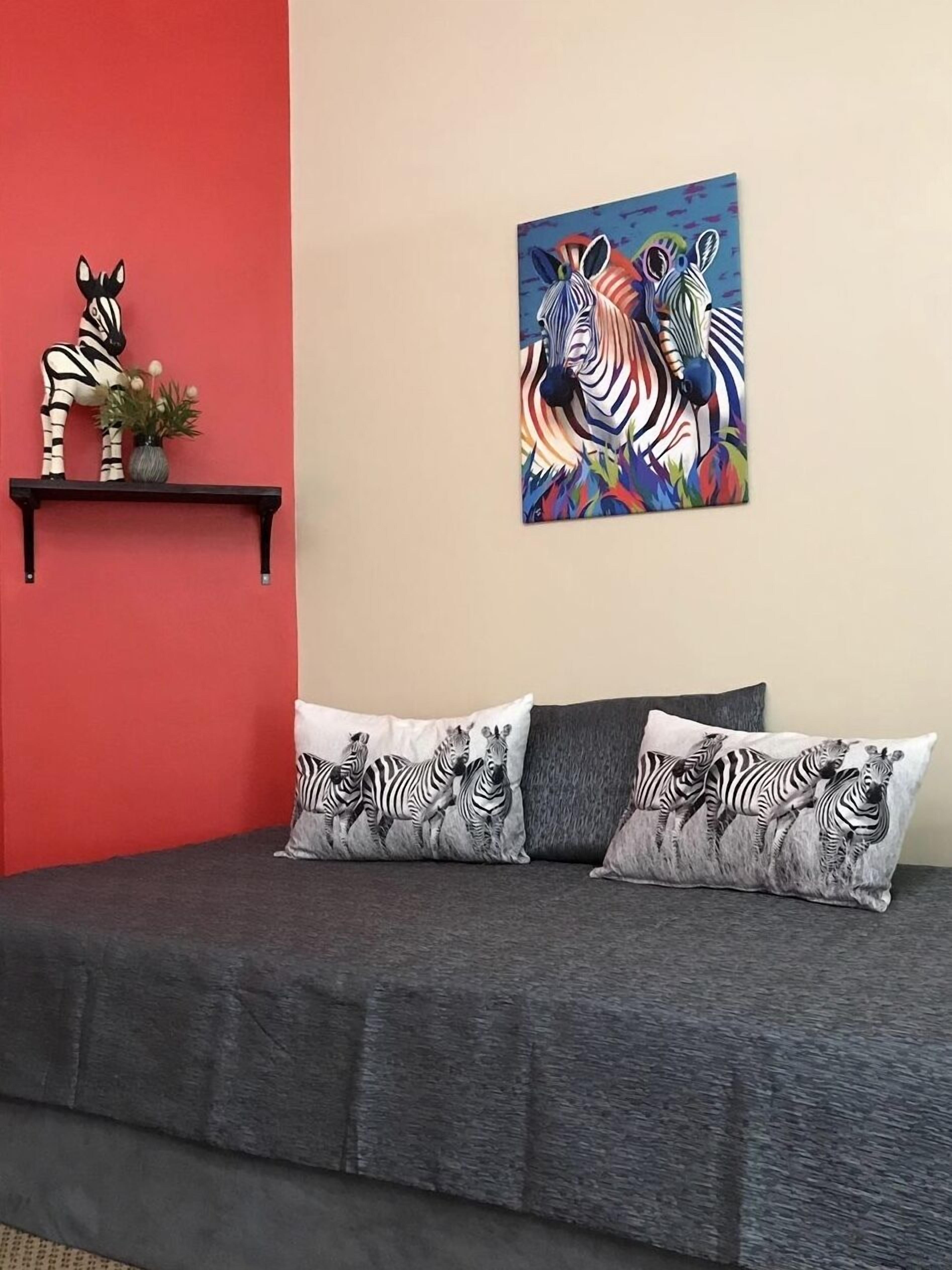 A bedroom with a grey sofa, zebra-themed pillows, a zebra statue, and a colorful zebra painting on the wall.