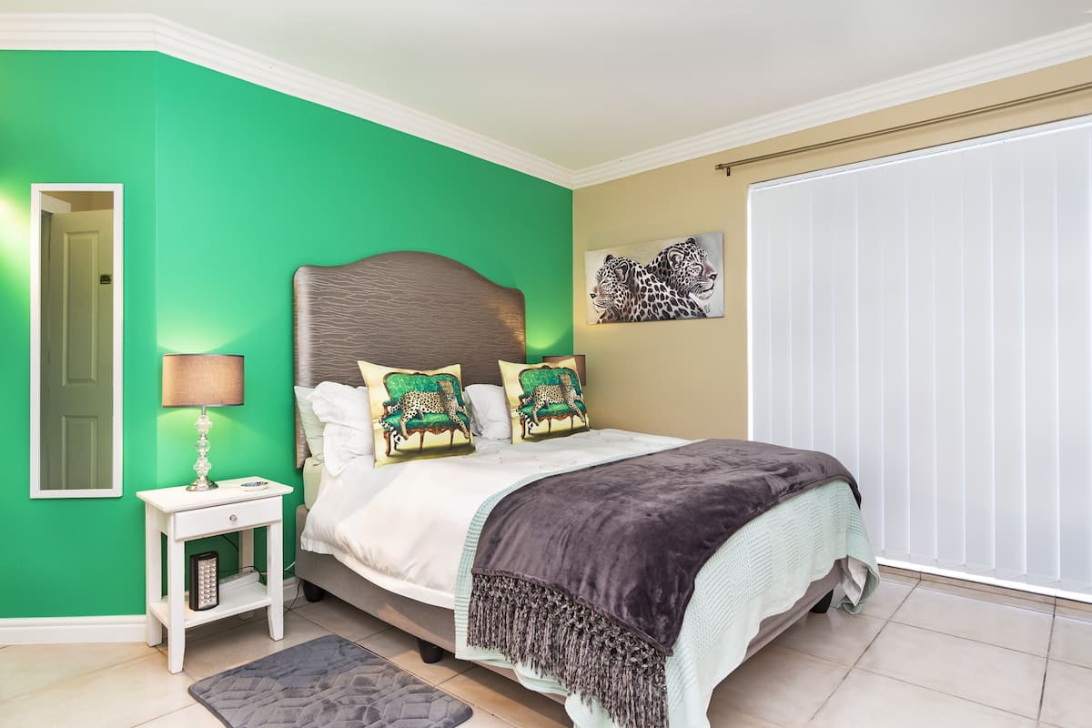 A bedroom with a green wall, a bed with a headboard, two decorative pillows, a bedside table with a lamp, and a window with blinds.
