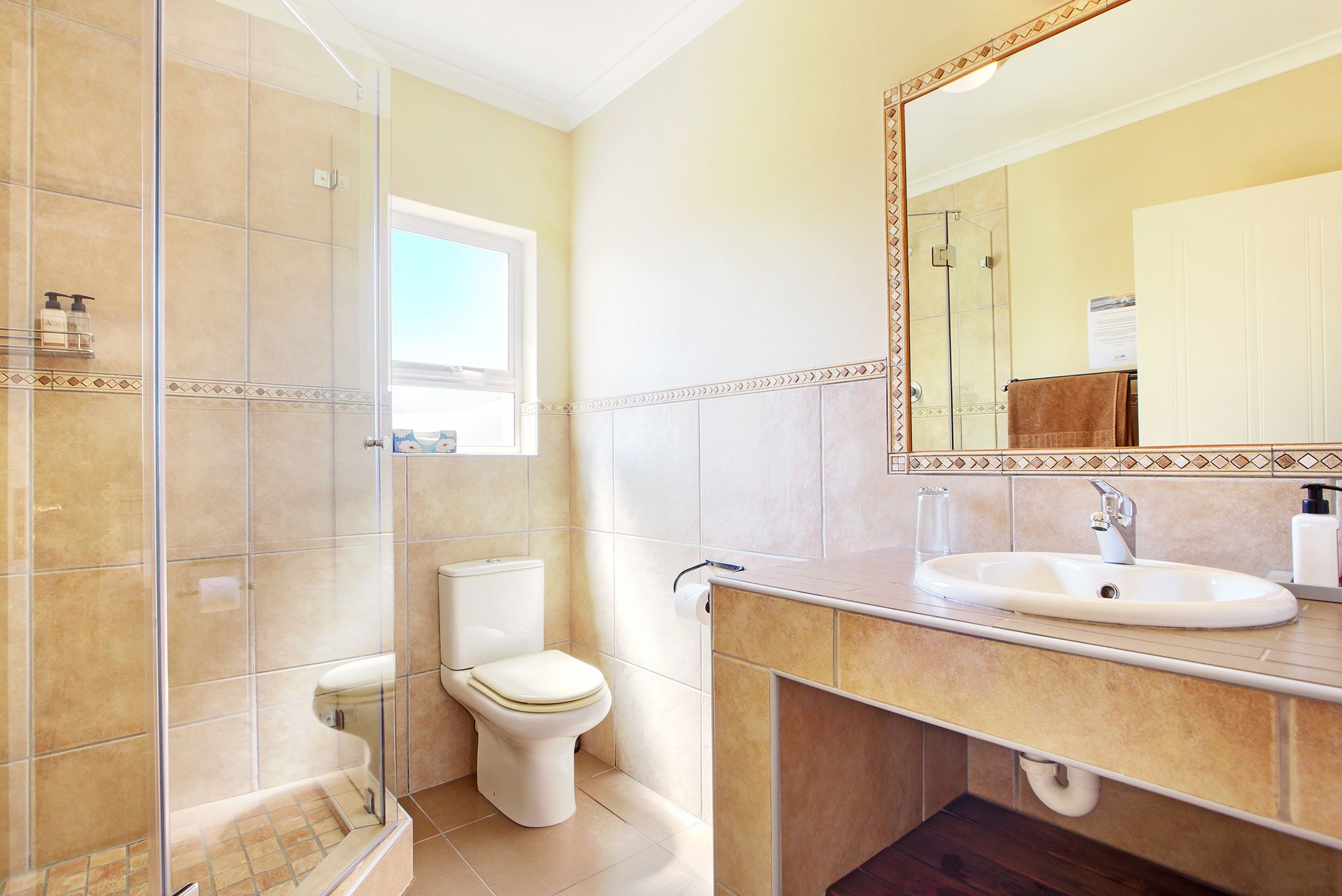 A bathroom with a glass shower enclosure, a toilet, a sink with a mirror, and a window providing natural light.