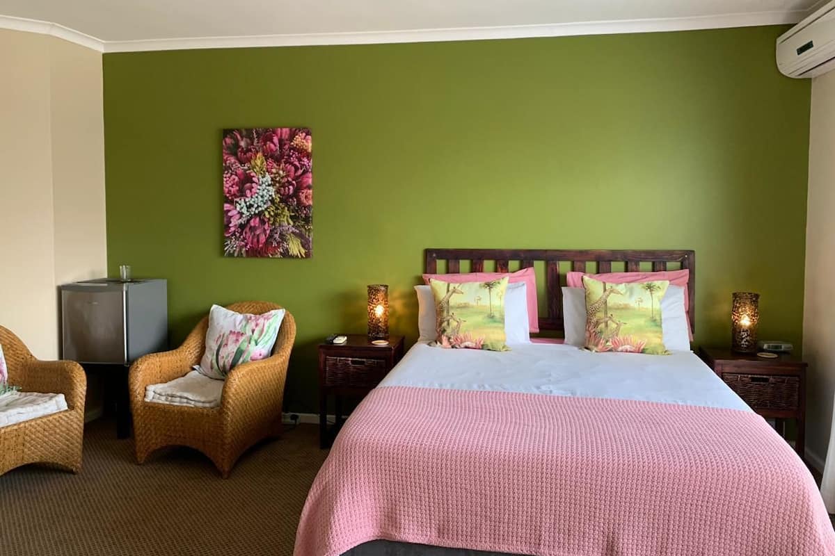 A hotel room with a large bed, two wicker chairs, a small table, a decorative wall piece, and a heater.