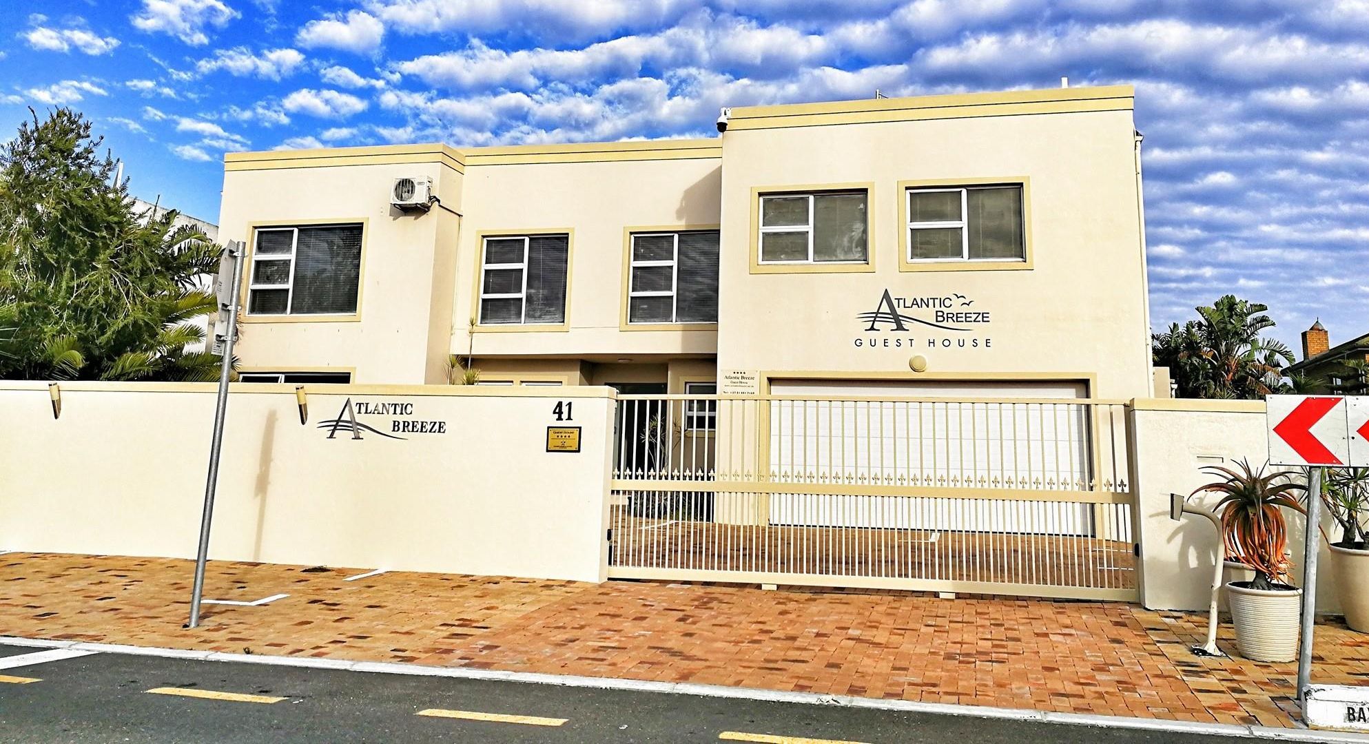 A two-story building with a beige facade, labeled "Atlantic Breeze Guest House," featuring a gated entrance, a number "41" sign, and a clear sky with clouds.