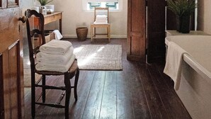 A room with a wooden floor, a wooden chair, a white bed, a window with shutters, a mirror, and a hanging light fixture.