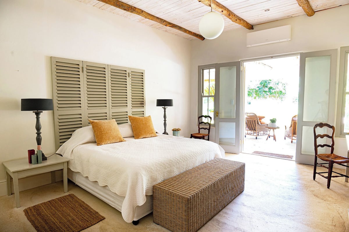 Un dormitorio con una cama, dos mesitas de noche con lámparas, un reposapiés de mimbre, una ventana con contraventanas y vistas a un patio exterior.