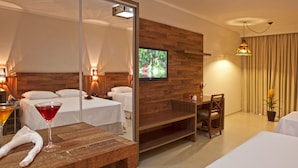 A hotel room with a wooden wall, a TV, a bed, a bedside table with a drink, and a chair. - Resort da Ilha (Sales)