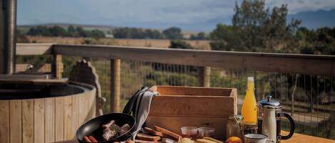 A wooden table with breakfast items, a kettle, and a bottle of orange juice on a balcony with a scenic view.