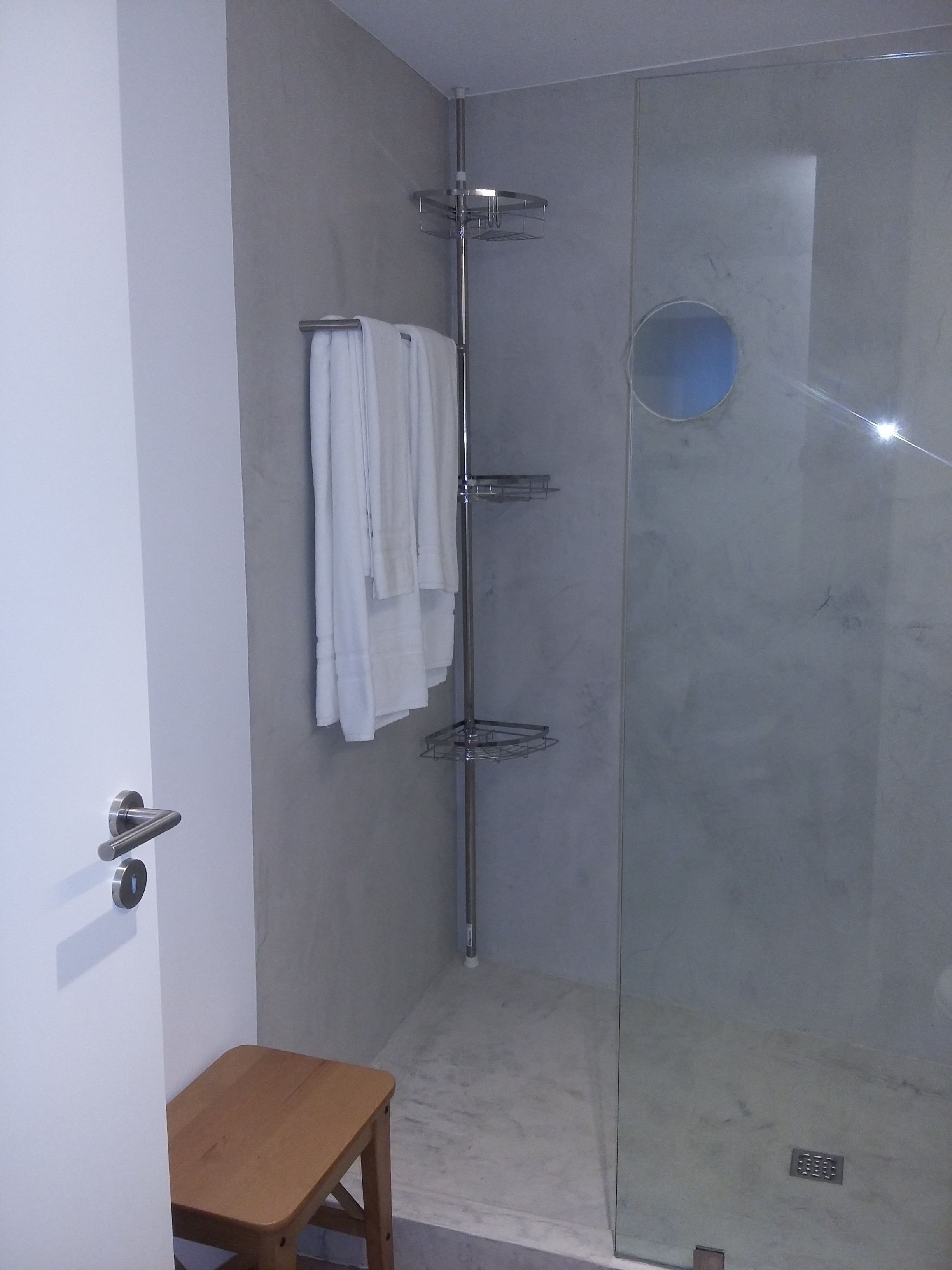 A modern bathroom with a glass shower enclosure, a towel rack with white towels, and a wooden stool.