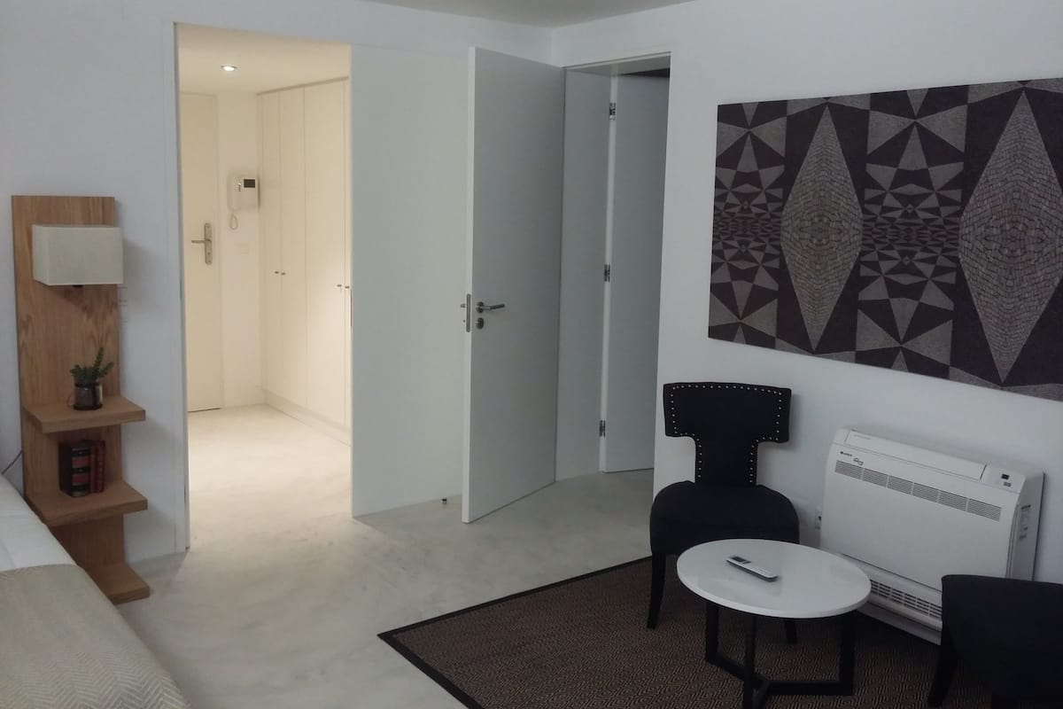 Une chambre d’hôtel moderne comprenant un lit, une petite table avec une chaise, un climatiseur fixé au mur et une œuvre d’art aux motifs géométriques.