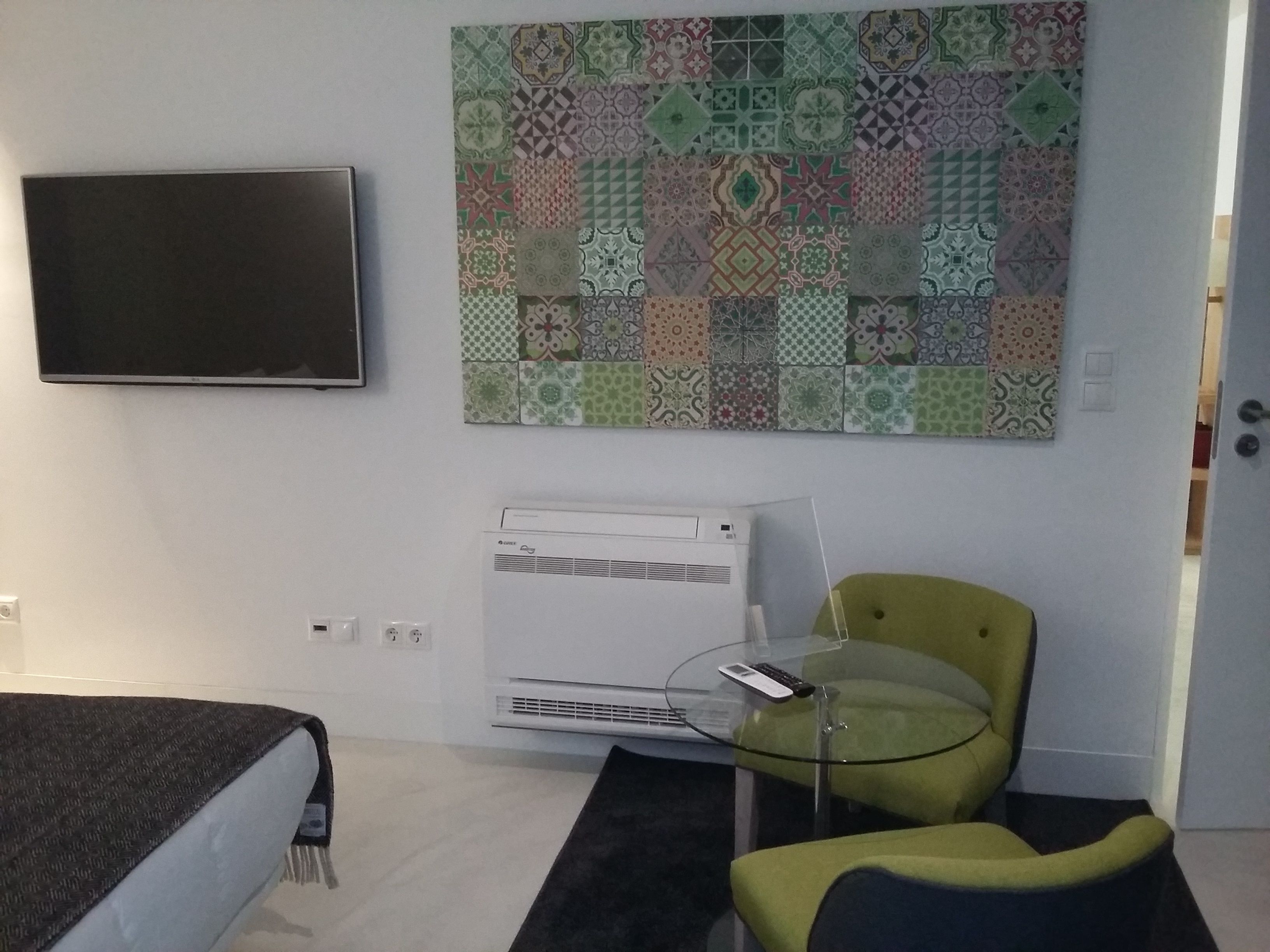 A modern hotel room with a flat-screen TV mounted on the wall, a patterned tapestry, a glass table with a remote control, a green armchair, and a bed with a dark cover.