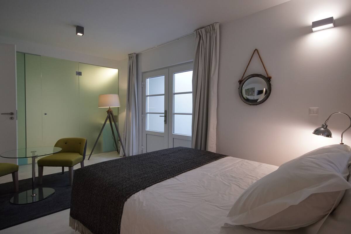 Une chambre d’hôtel comprenant un lit, un bureau avec une chaise, une lampe, un miroir et une fenêtre avec des rideaux.
