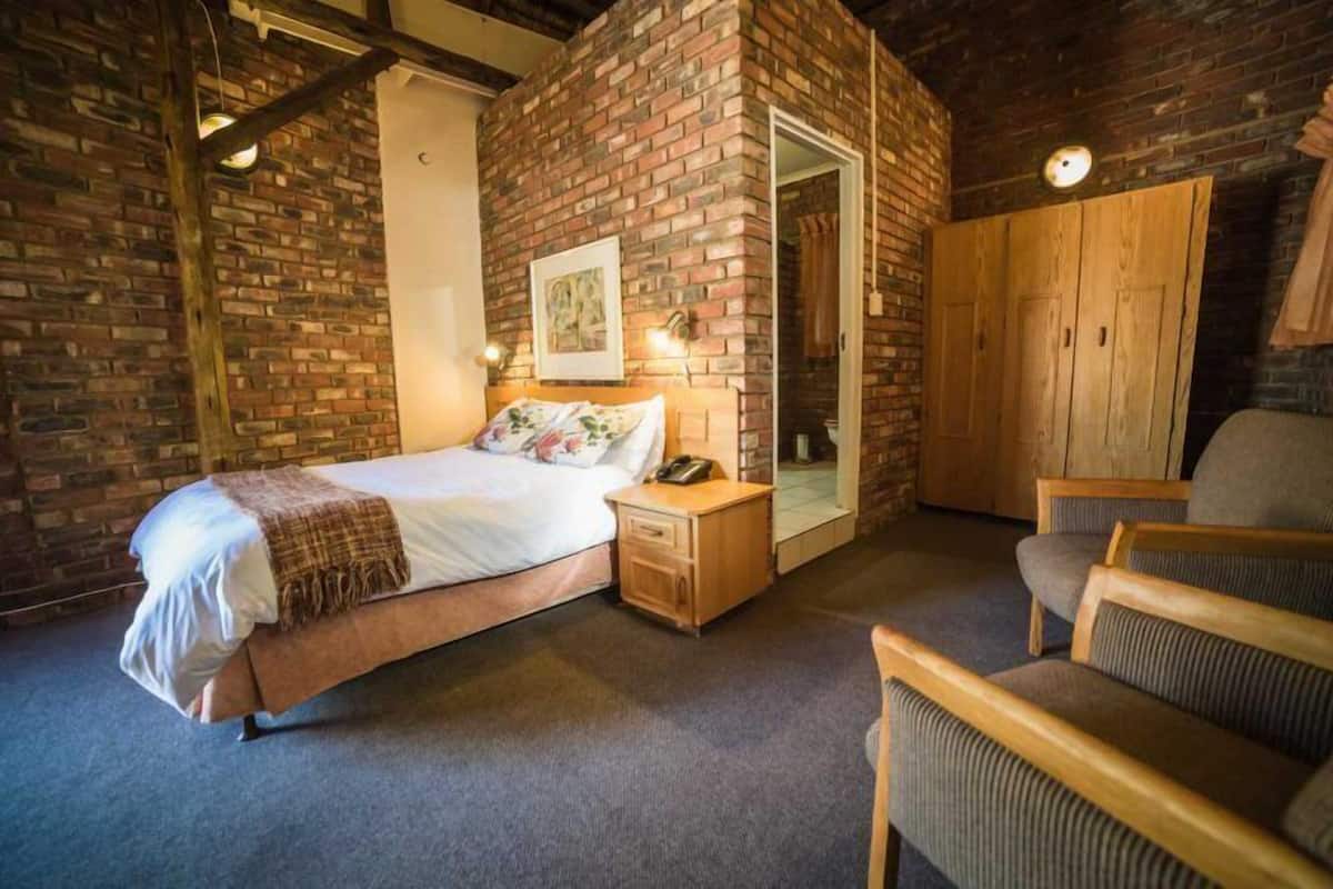 A bedroom with a brick wall, a bed with a white comforter, a wooden nightstand, a chair, and a wardrobe.