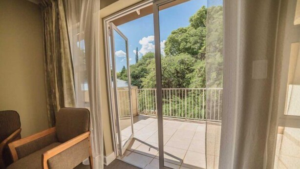 A room with a sliding glass door leading to a balcony with a view of trees and a building. - Acacia Guesthouse (Klerksdorp)