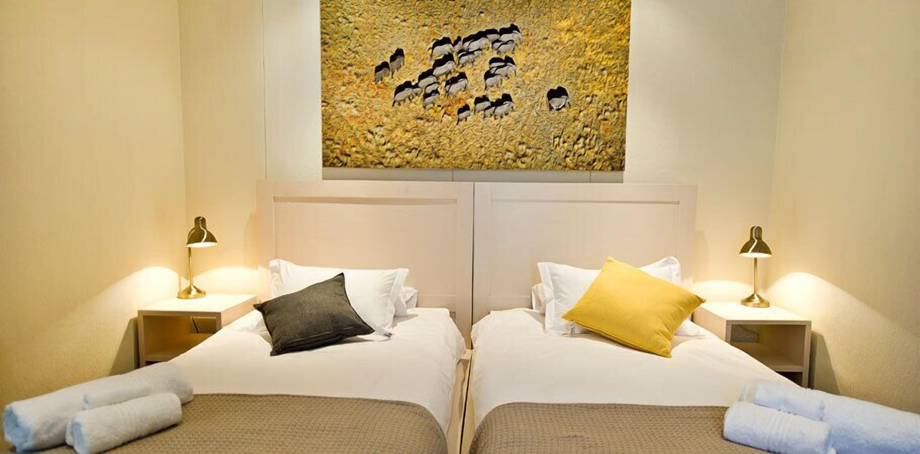 A hotel room with two beds, each with a pillow and a lamp. There is a painting above the beds.