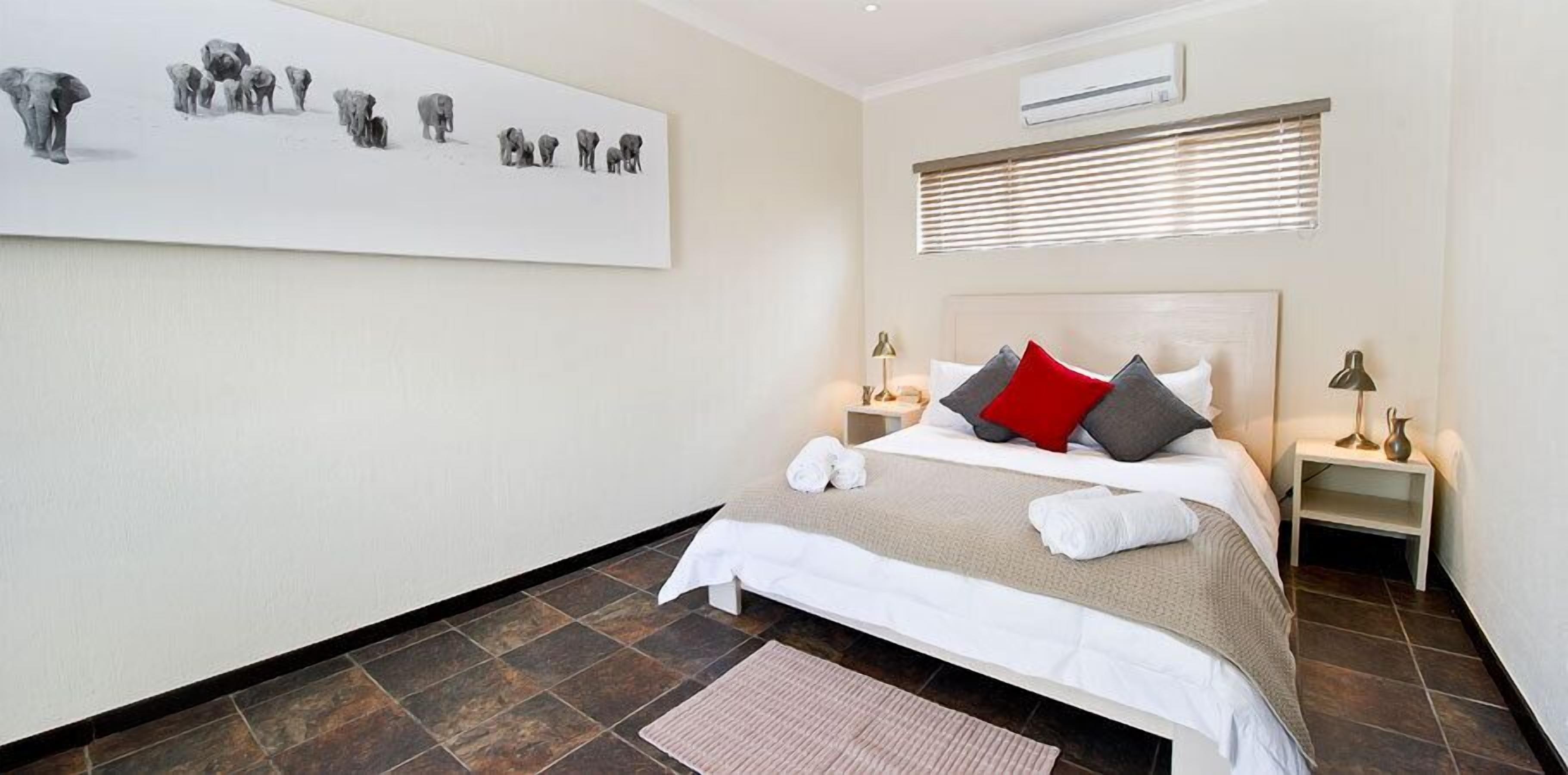 A hotel room with a large bed, two bedside tables with lamps, a wall-mounted air conditioner, and a large painting of elephants.