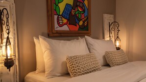Un lit avec du linge de lit blanc, deux coussins à motifs, une peinture abstraite colorée au-dessus du lit et deux lampes murales de style lanterne.