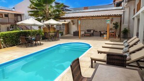 Une piscine avec des chaises longues, un espace salon couvert et une terrasse avec des tables et des parasols.