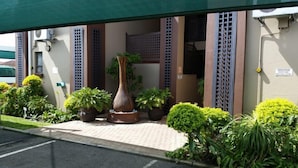 Une entrée d’immeuble avec un chemin bordé de carreaux, des plantes en pot et un grand vase.