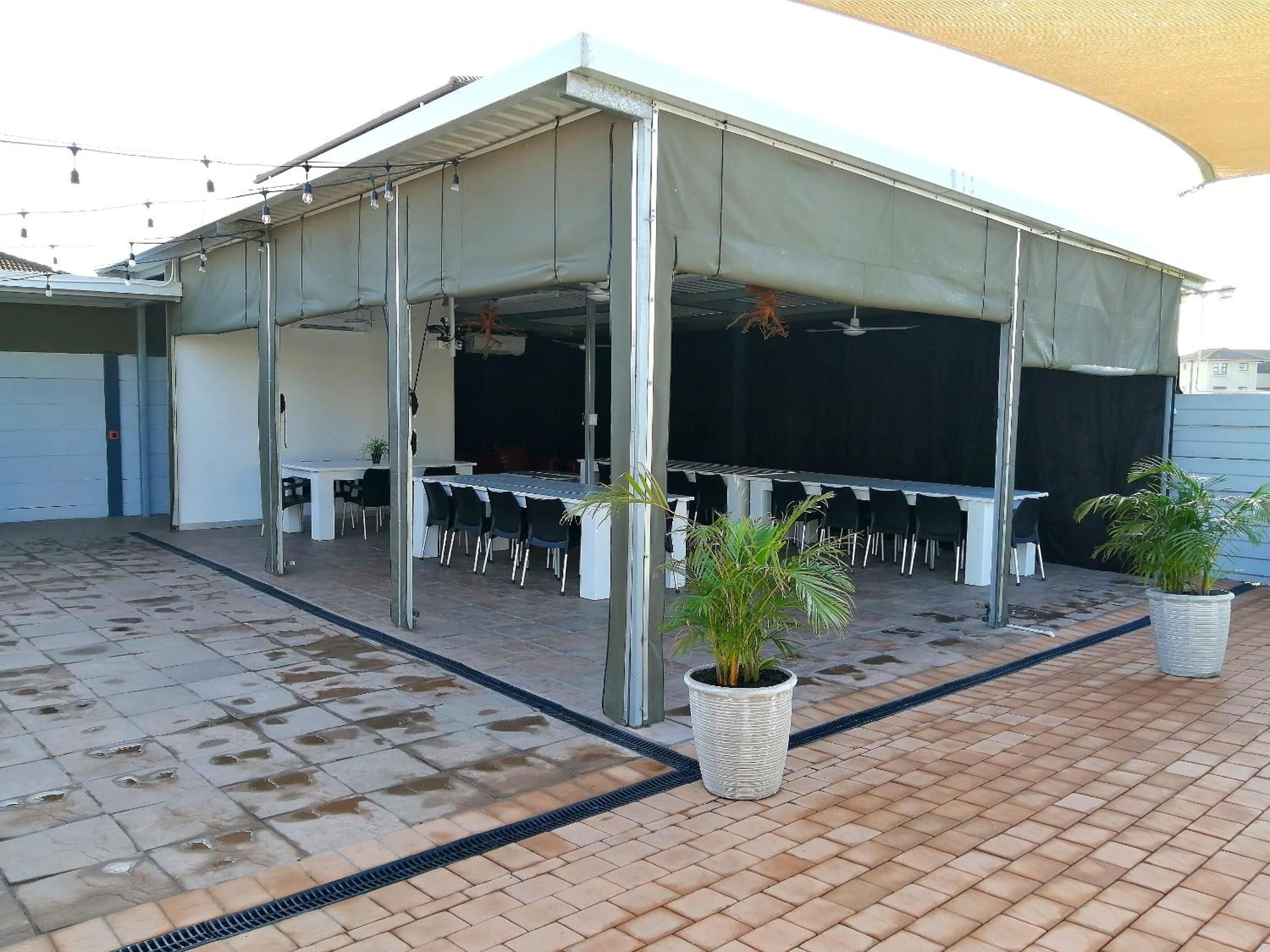 Un'area patio esterna coperta, con tavoli e sedie, una ventola a soffitto e piante in vaso.