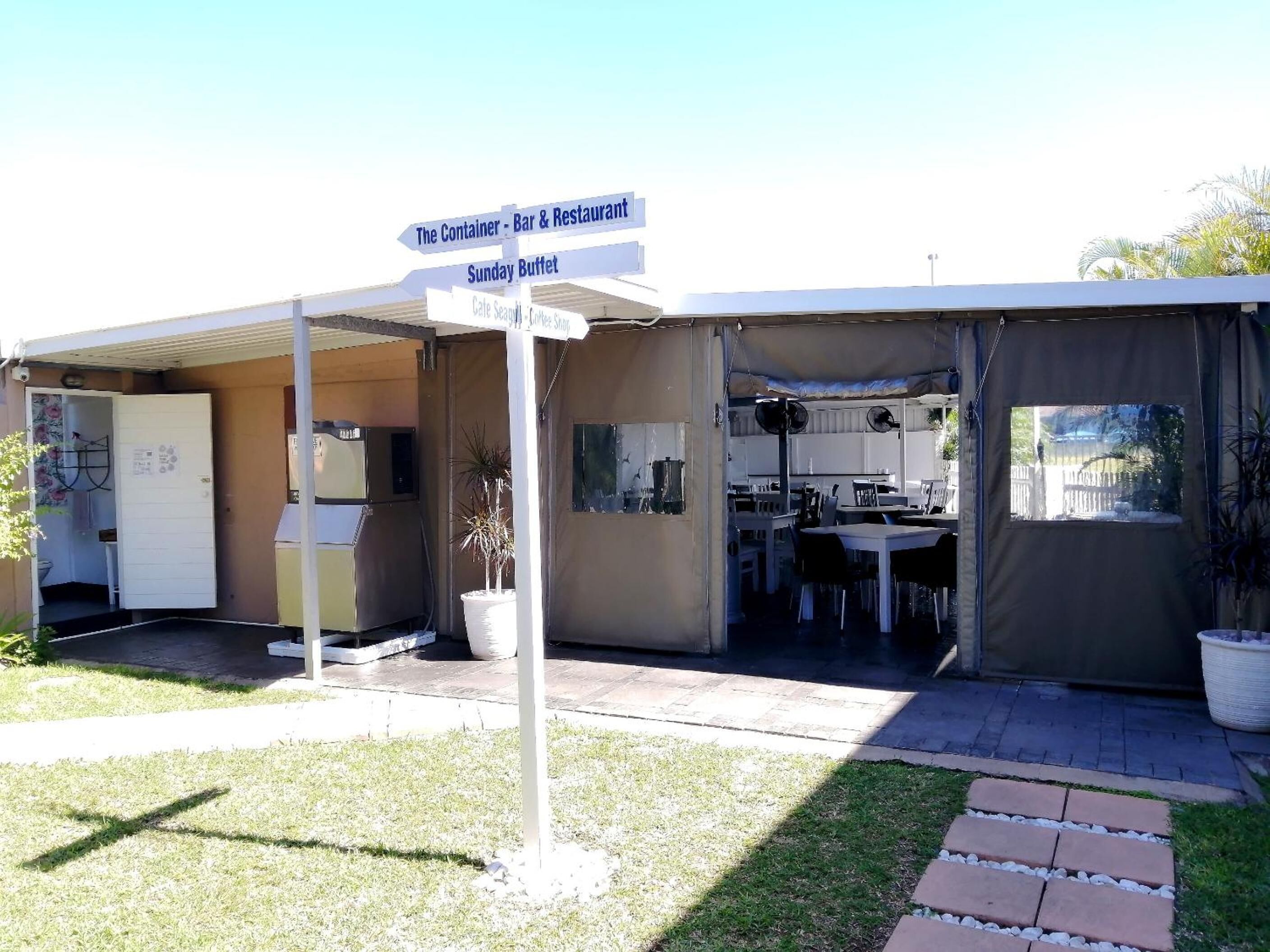 A small, single-story building with a covered outdoor area. There is a sign indicating 'The Container - Bar & Restaurant' and 'Sunday Buffet'. Inside, there are tables and chairs arranged for dining.