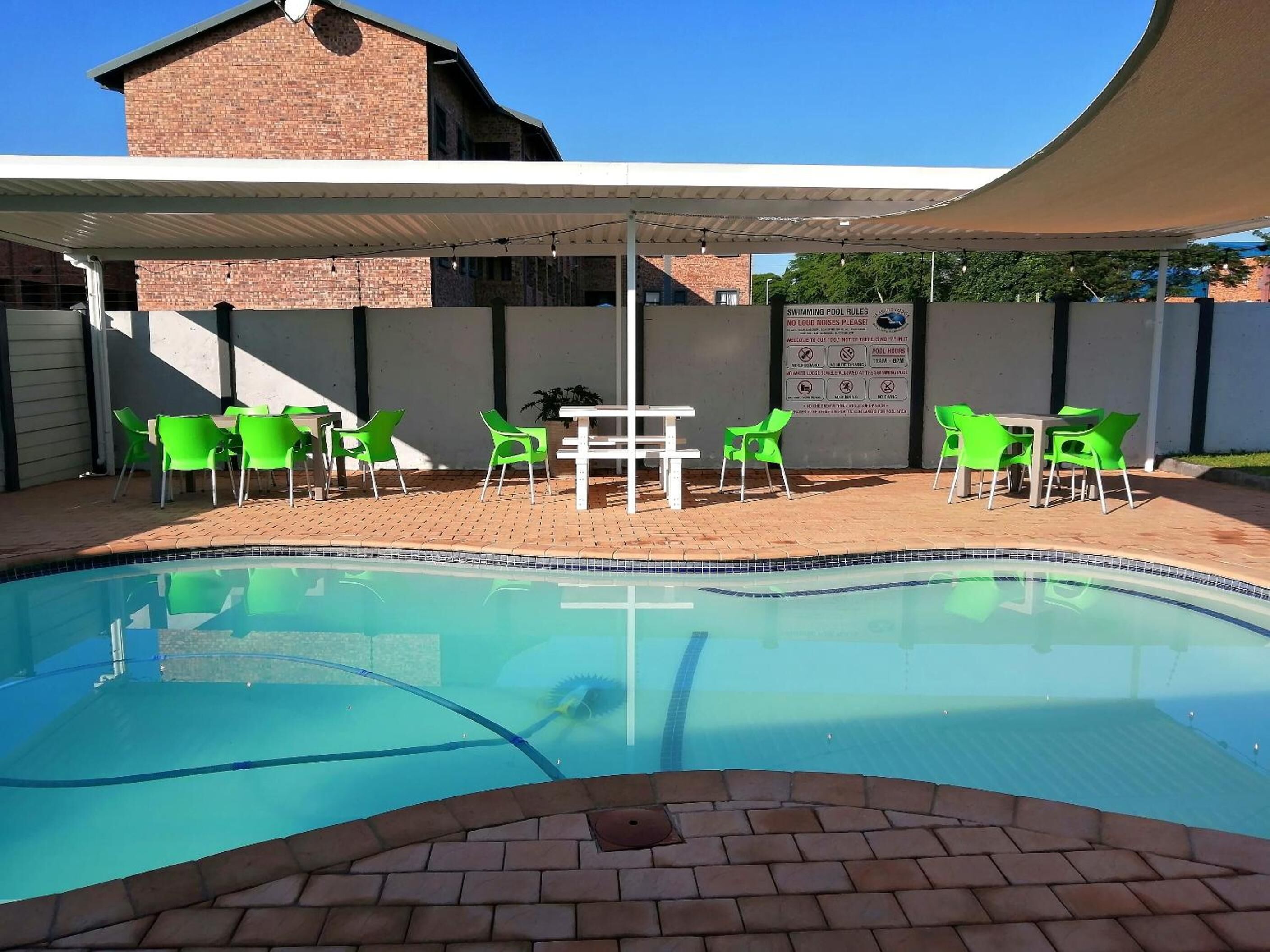 A swimming pool with a covered patio area, green chairs, and a sign with pool rules.