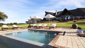 A pool area with lounge chairs and umbrellas, a large house with a sloped roof, and a grassy lawn. - JBay Zebra Lodge (Thornhill)