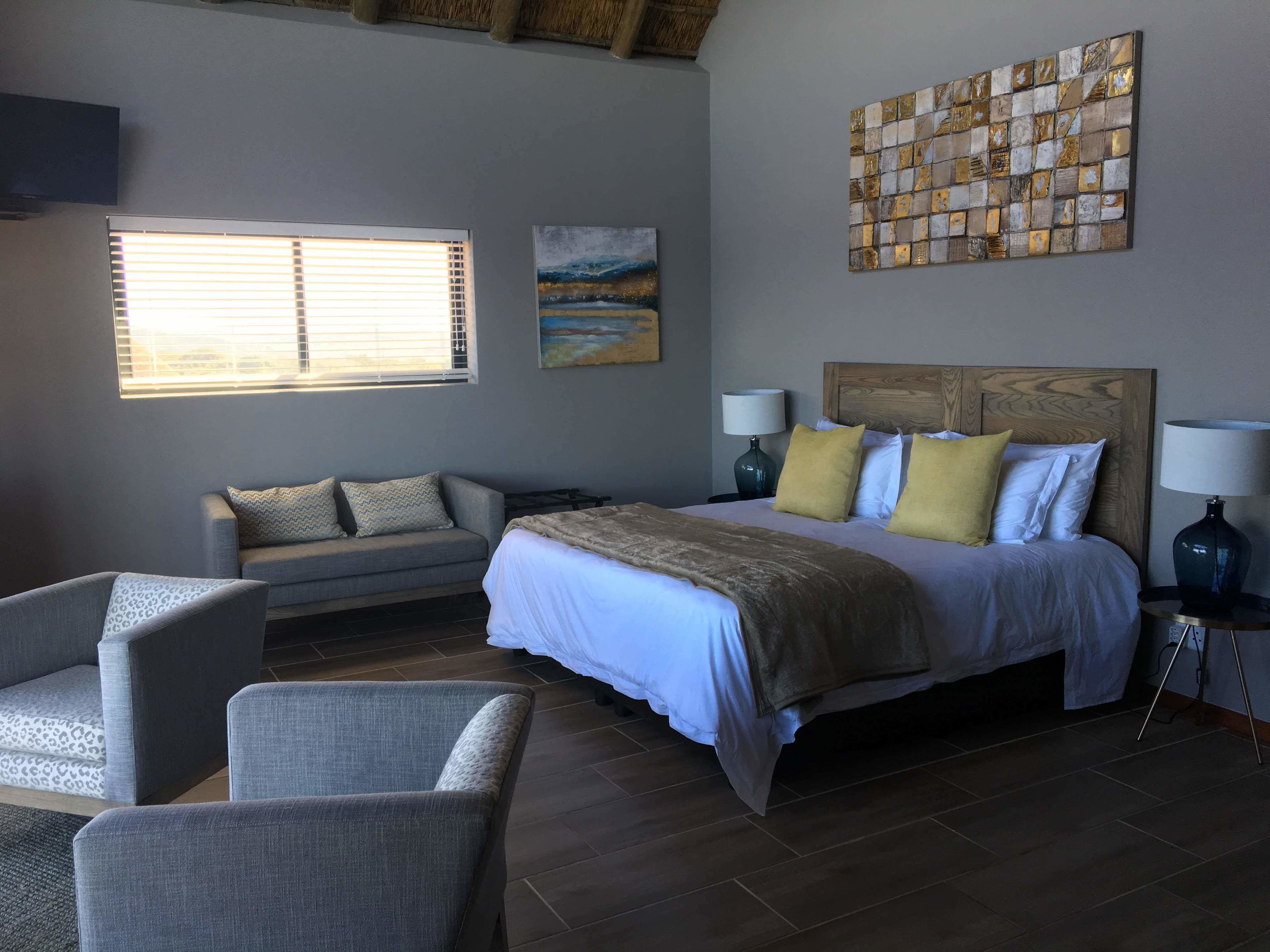 A hotel room with a bed, a sofa, two armchairs, a painting, and a window with blinds.