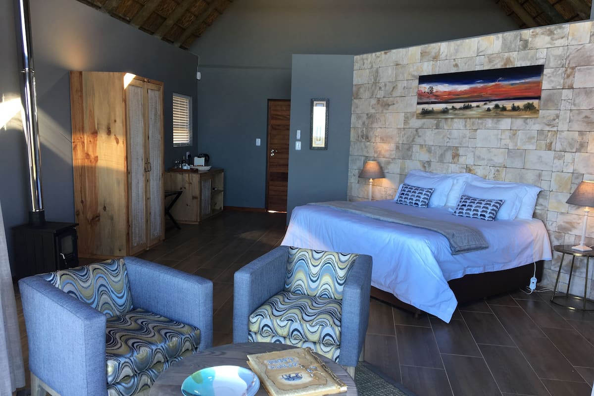 A bedroom with a bed, two armchairs, a table, a fireplace, and a stone wall with a painting.