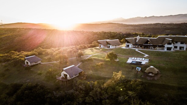 An aerial view of a resort with multiple buildings, including a large house with a unique roof design, smaller cottages, and a swimming pool area. - JBay Zebra Lodge (Thornhill)