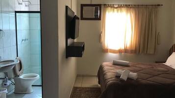 Habitación de hotel con cama, ventana con cortinas, televisor montado, zona de ducha y un inodoro.
