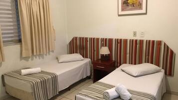 A hotel room with two single beds, a striped headboard, a bedside table with a lamp, a window with curtains, and a framed picture on the wall.