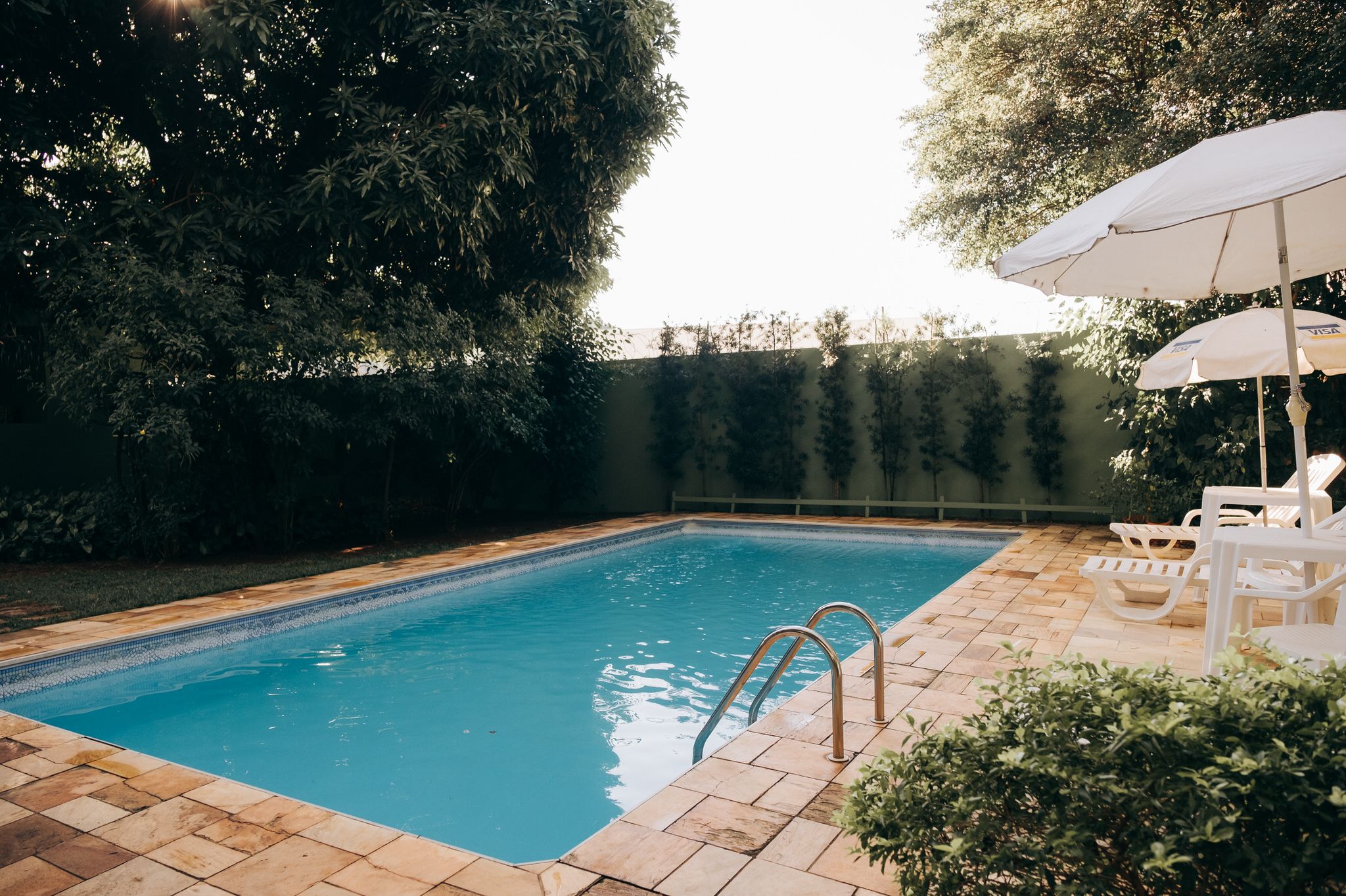 A rectangular swimming pool with a tiled edge, surrounded by a brick patio. There are two white lounge chairs and a white umbrella on the patio.