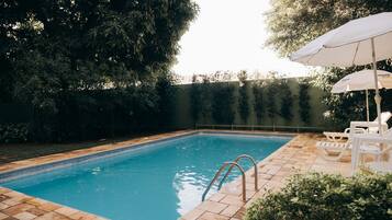 A rectangular swimming pool with a tiled edge, surrounded by a brick patio. There are two white lounge chairs and a white umbrella on the patio.