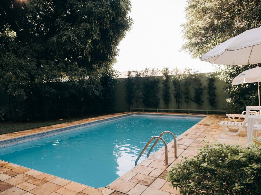 A rectangular swimming pool with a tiled edge, surrounded by a brick patio. There are two white lounge chairs and a white umbrella on the patio.