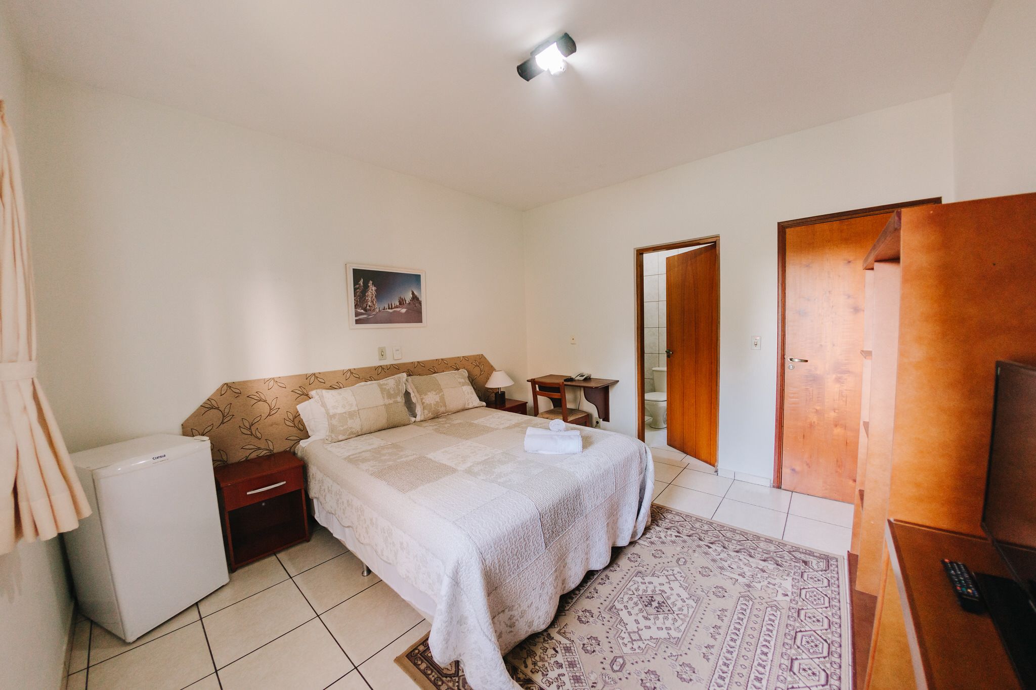 A bedroom with a bed, a small refrigerator, a bedside table with a lamp, a wooden door, and a television.