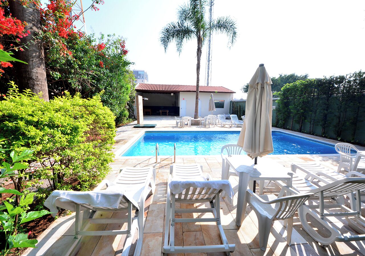 A poolside area with lounge chairs and an umbrella, surrounded by greenery and a building with a red roof.
