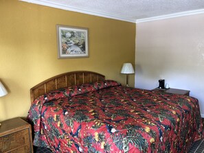 A room with a wooden headboard bed, a floral bedspread, a framed picture on the wall, a bedside table with a lamp, and a nightstand with a coffee maker.