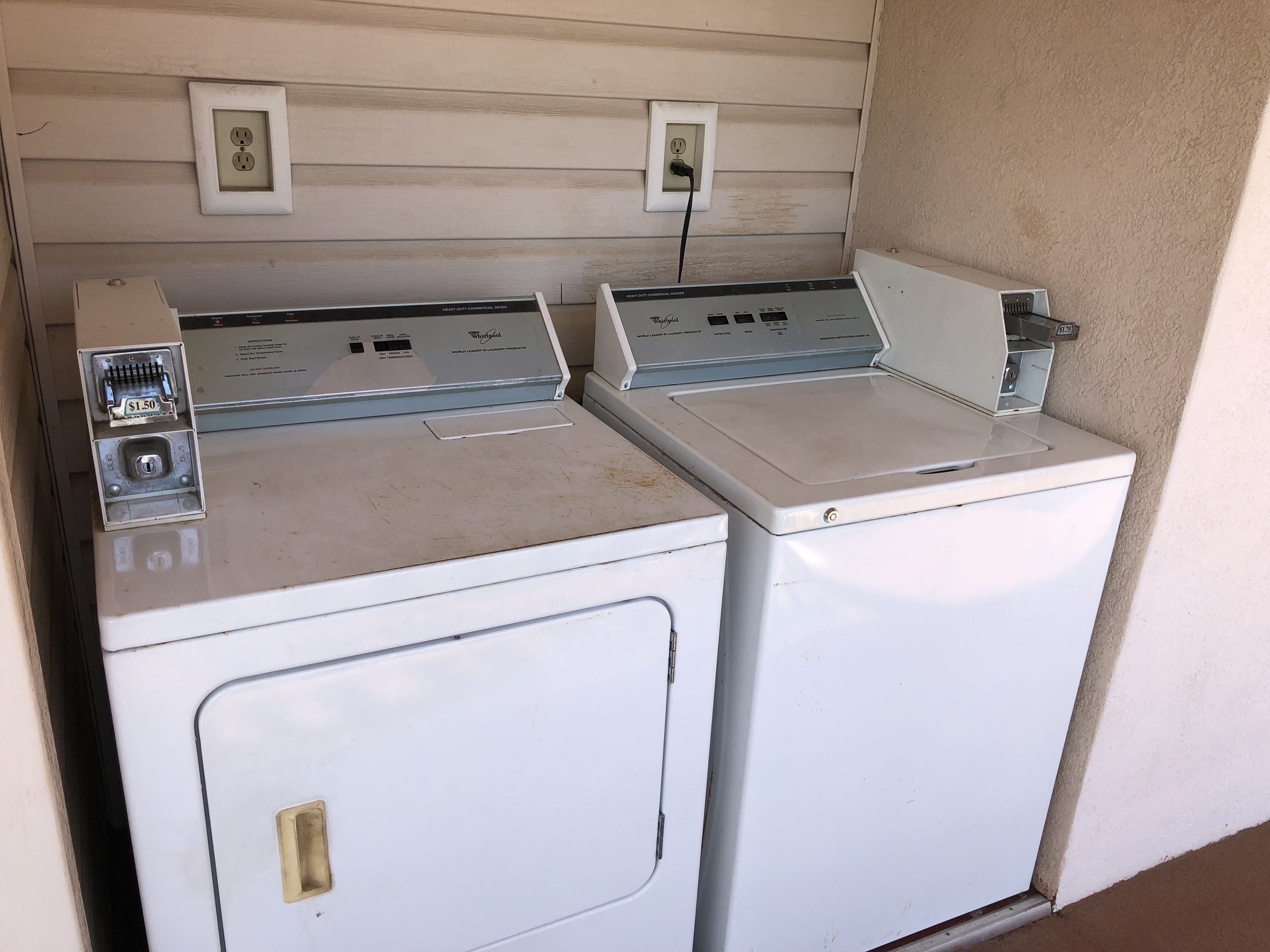Two stacked front-loading washers with control panels, a coin slot, and a wall-mounted electrical outlet.