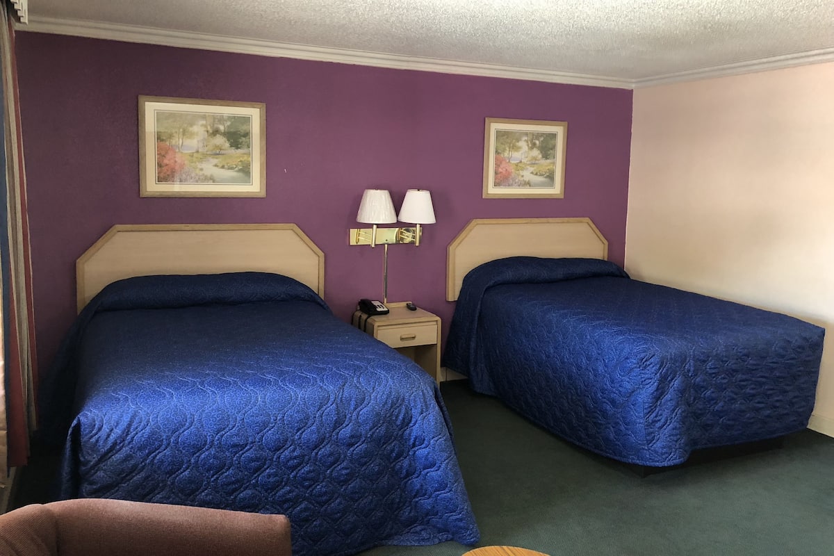 Hotellihuone, jossa on kaksi sänkyä, violetit seinät ja kaksi kehystettyä kuvaa.