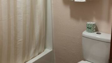 A bathroom with a shower curtain, a toilet with a paper towel holder, and a shelf with towels.